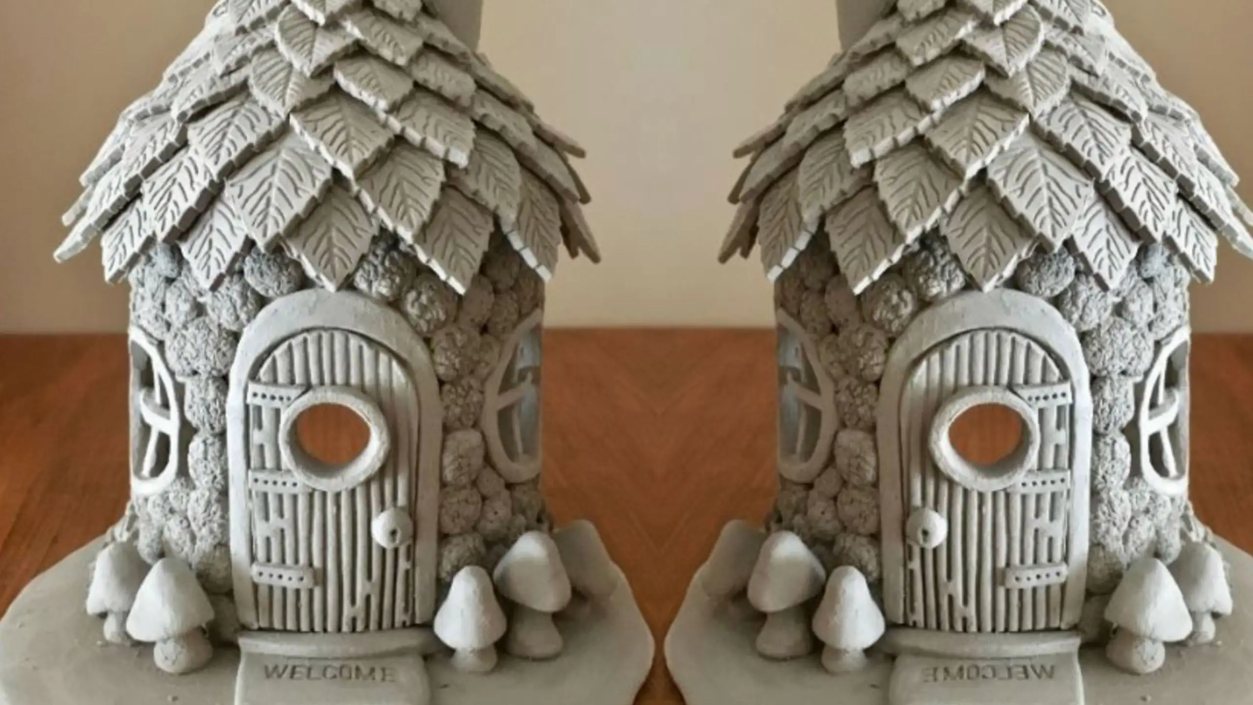 Two handcrafted clay fairy houses with leaf-patterned roofs, textured stone walls, arched doors and tiny mushrooms, displayed on a wooden surface.